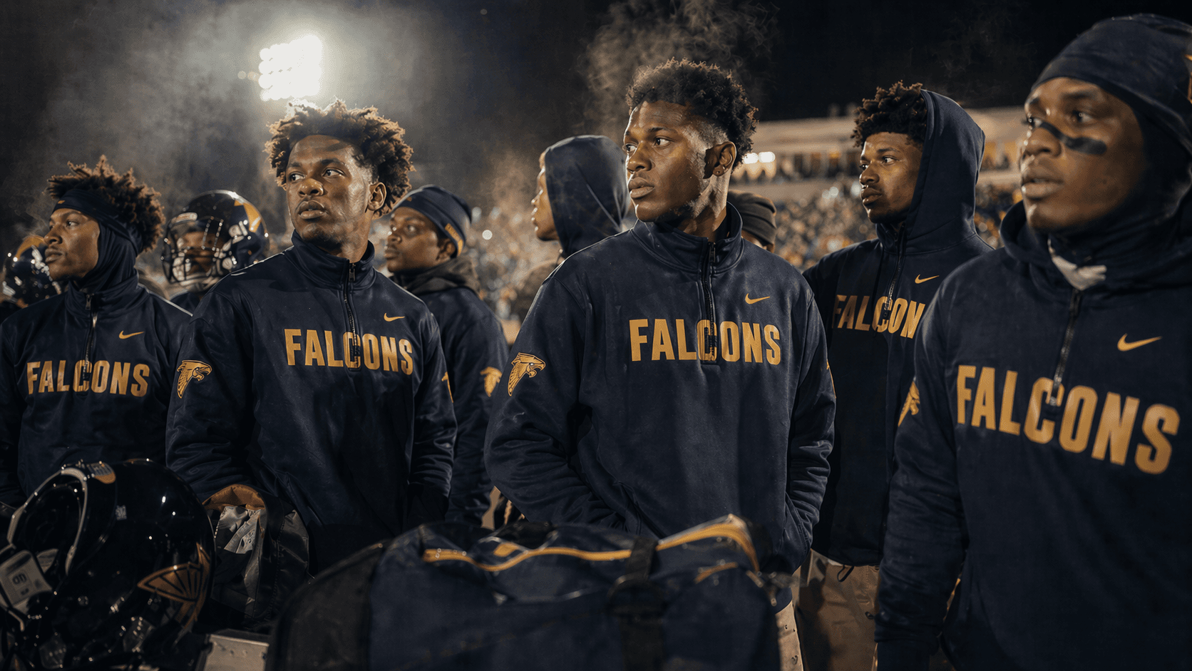 Football sideline under stadium lights, players in navy quarter-zips with a printed gold wordmark across the chest and an embroidered crest on the shoulder.