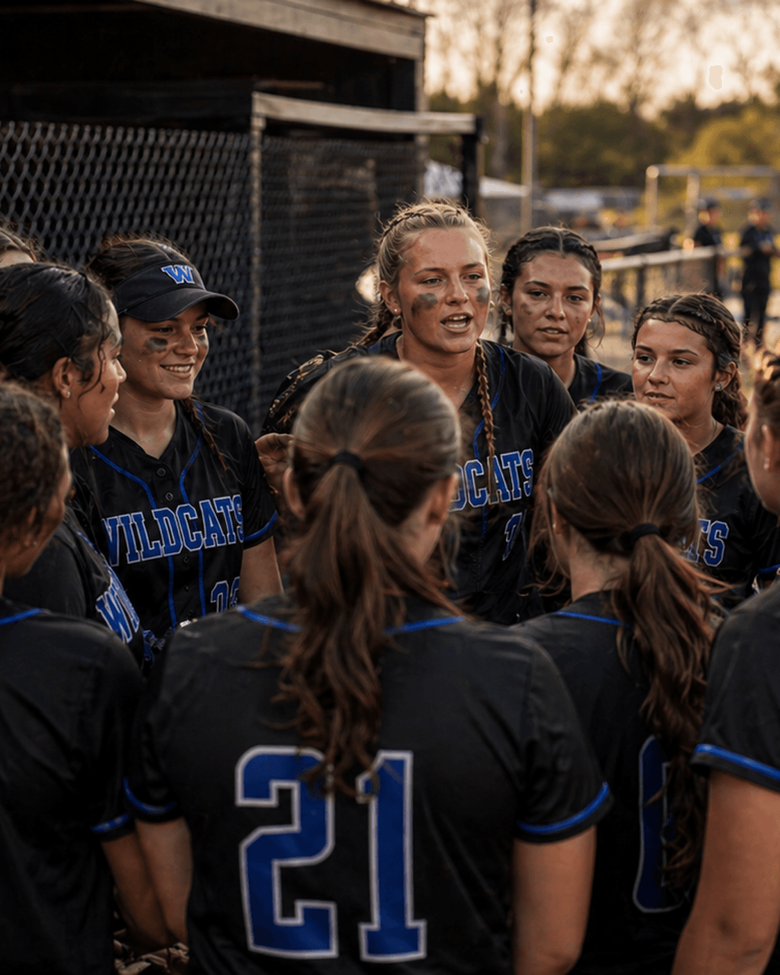High school fastpitch softball team huddled outside the dugout at golden hour in black jerseys with tackle twill chest lettering and blue contrast piping.