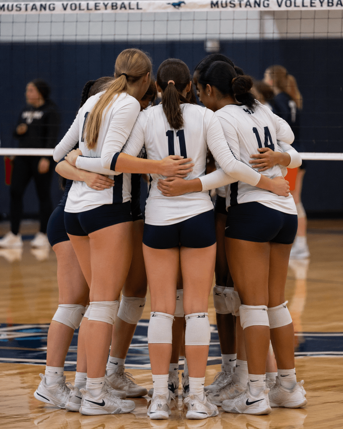 Girls high school volleyball team huddled at center court between points, white jerseys with navy shorts.